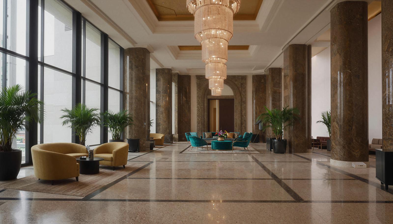A spacious luxury hotel lobby with marble columns, a chandelier hanging from the ceiling, sofas and houseplants arranged throughout
