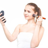 コンパクトミラーを持ちながらチークブラシでメイクする外国人女性の写真