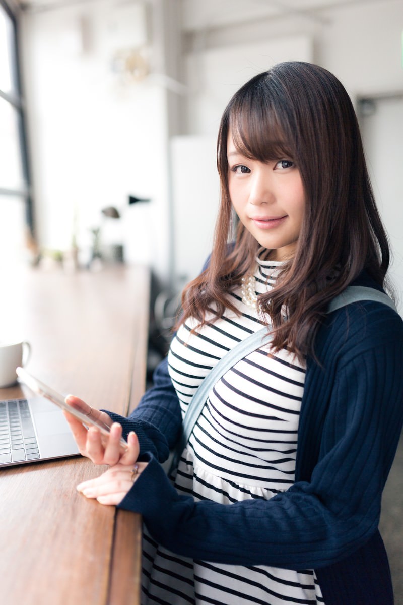 スマートフォンを握りながら笑顔でカメラを見つめるストライプシャツの女性［モデル：茜さや］