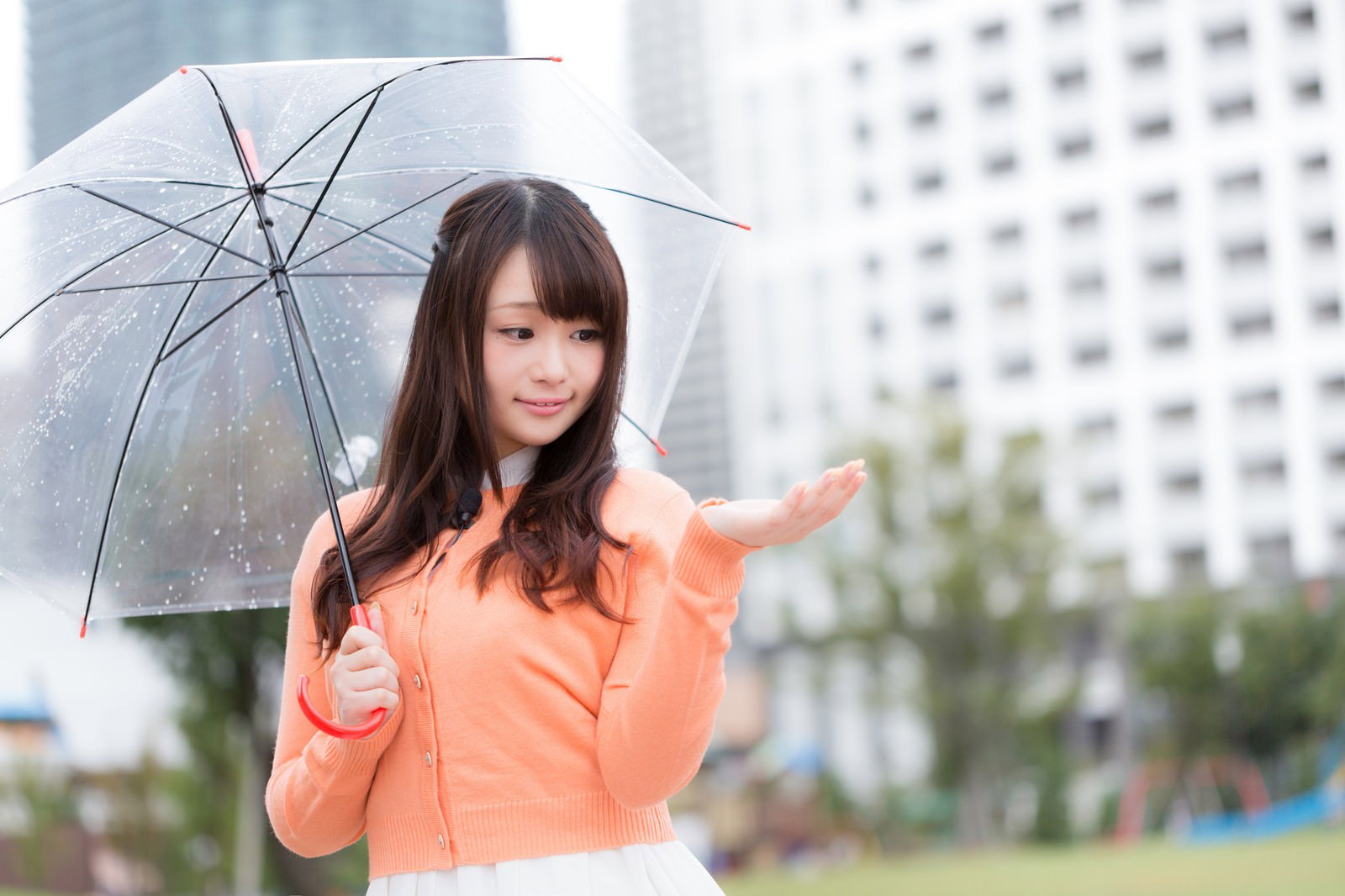 オレンジのシャツを着た女性が傘を持ち、雨の中で立つ写真［モデル：茜さや］