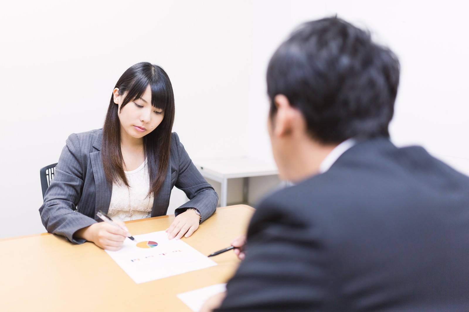 グレースーツの女性がテーブルのグラフ資料を指しながら、黒いスーツの男性に説明している［モデル：大川竜弥 Lala］