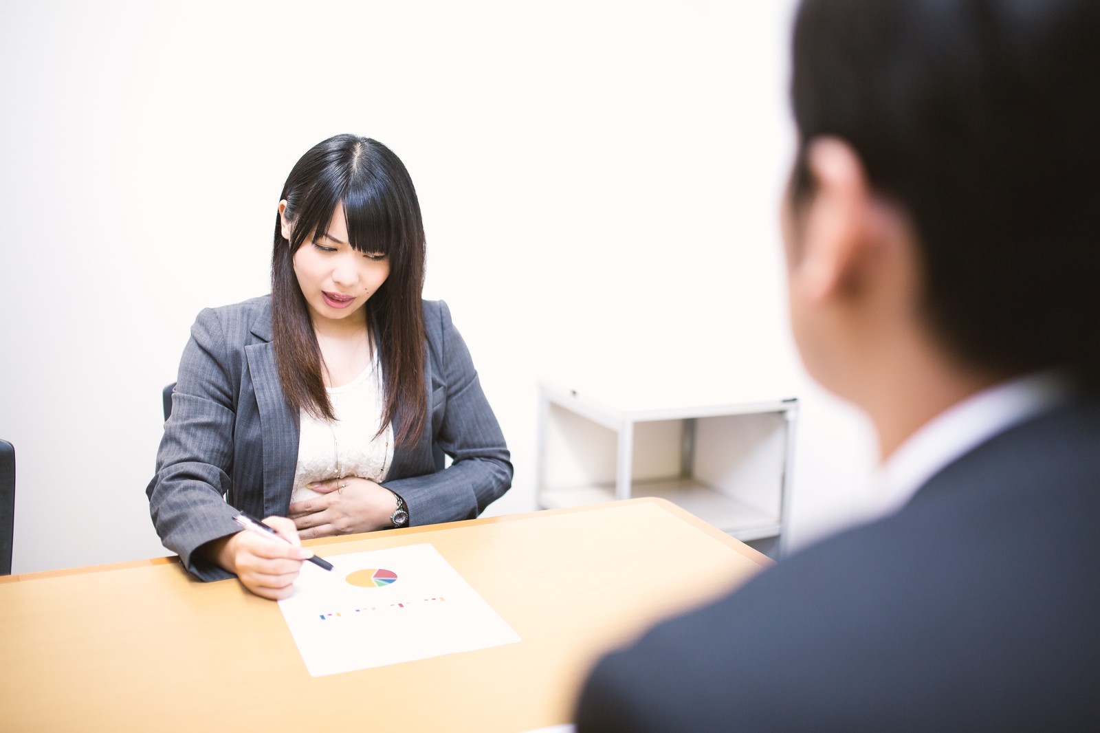 会議中にお腹を押さえるスーツ姿の女性［モデル：大川竜弥 Lala］