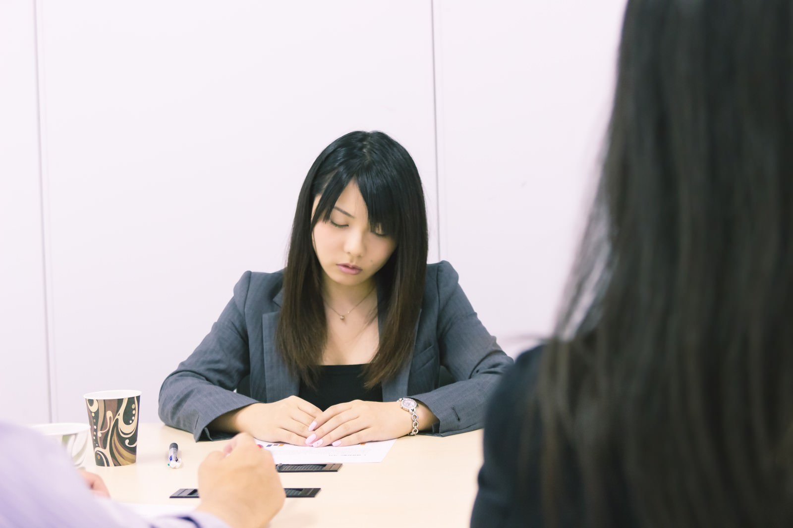 机で書き物をする女性と対面のぼけた人物の会議シーン［モデル：Lala］