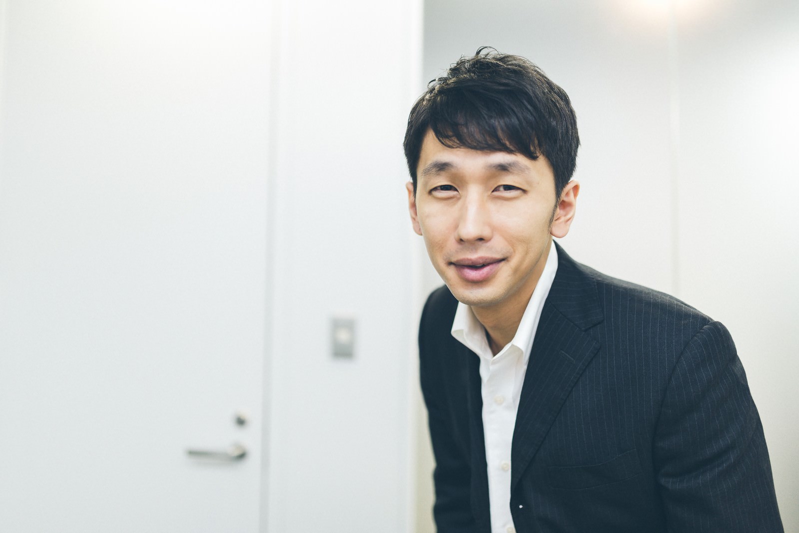 黒いストライプスーツとワイシャツを着た男性が不適切な笑顔を浮かべる室内ポートレート［モデル：大川竜弥］