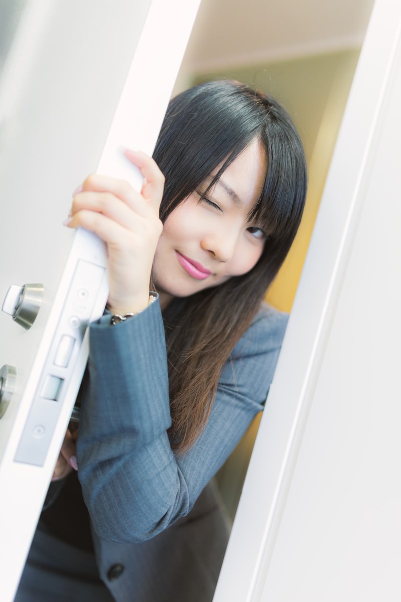 紺色スーツを着た女性がドアから顔を出し、ウインクしながら笑顔を見せている［モデル：Lala］