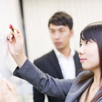 ホワイトボードに書き込む女性社員と見守る男性の写真