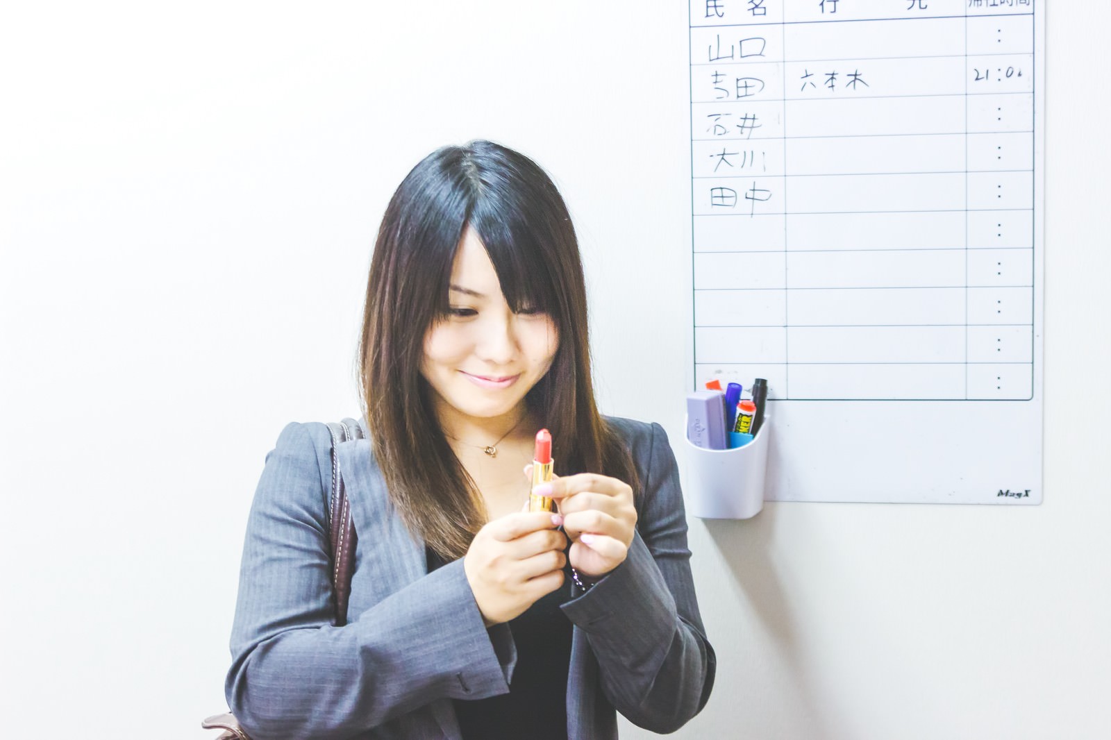 紺色スーツの女性が赤い口紅を両手で握り、手元を見つめている［モデル：Lala］