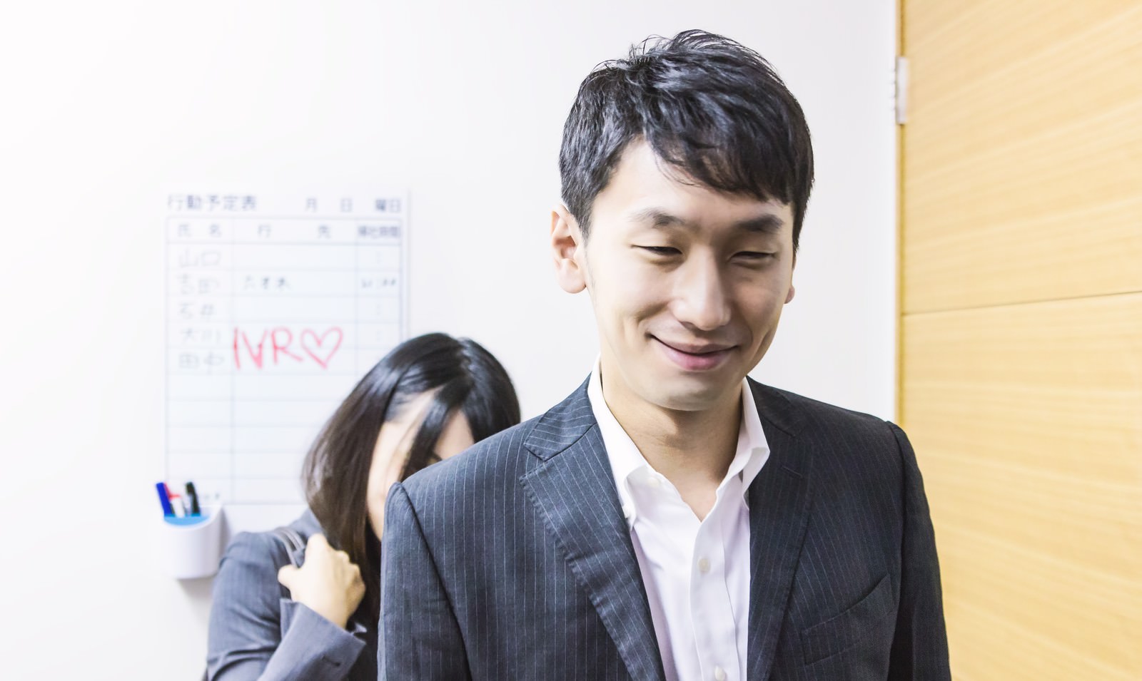 紺ストライプスーツの男性が目を閉じて微笑み、背後に予定表を持つ女性社員が立つオフィスシーン［モデル：大川竜弥 Lala］