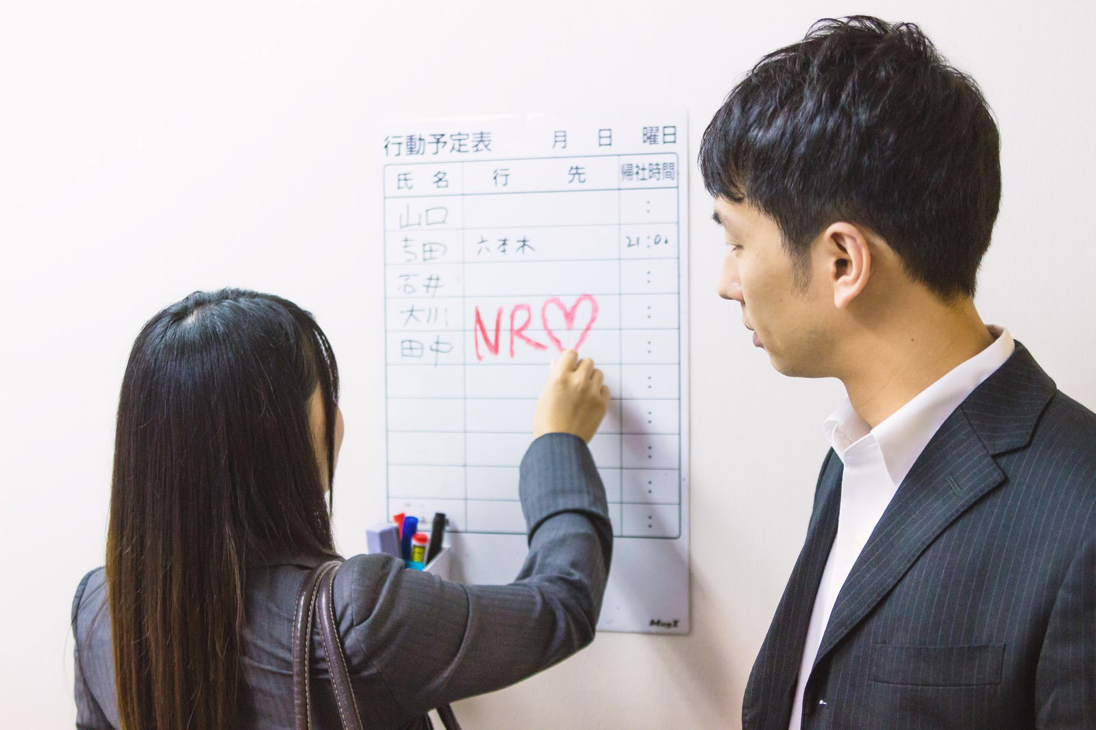 行動予定表にNR♡と赤で書き込む女性社員と、その様子を見守る男性社員［モデル：大川竜弥 Lala］