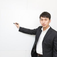 白い壁をペンで指しながらプレゼンする黒ストライプスーツの男性の写真