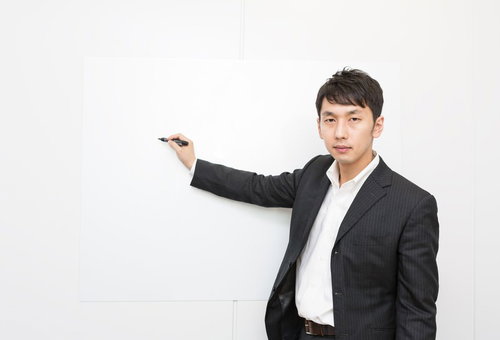 白い壁をペンで指しながらプレゼンする黒ストライプスーツの男性