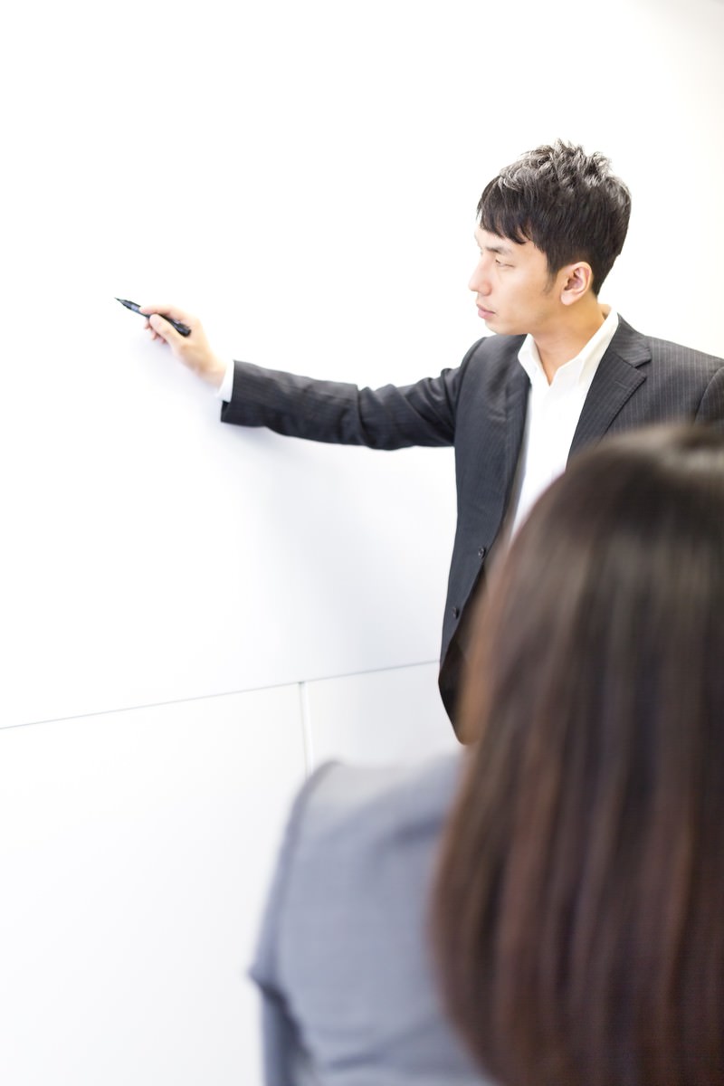 ホワイトボードにペンで書きながら説明するスーツ姿の男性［モデル：大川竜弥 Lala］