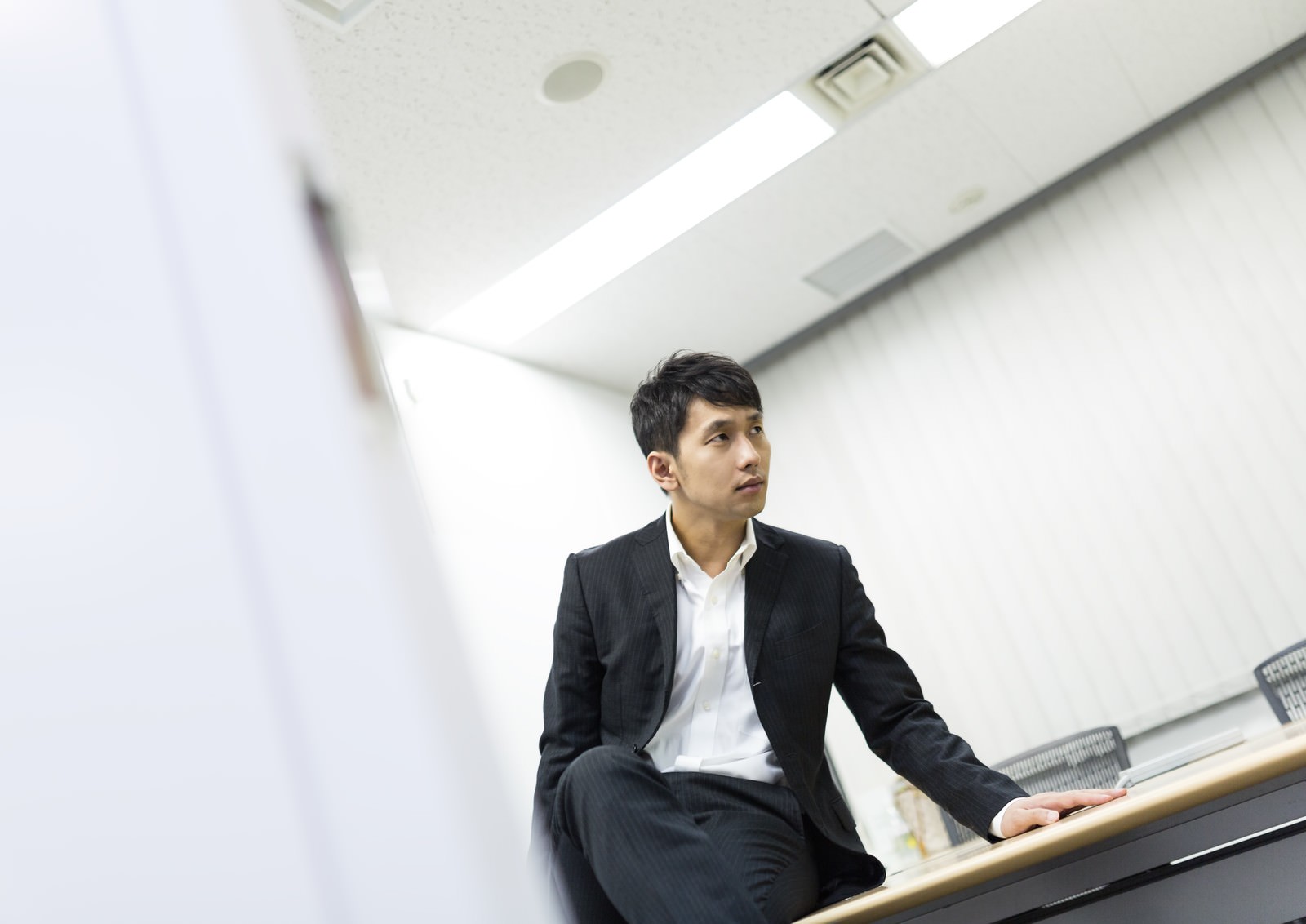 机に腰かけて斜め上を見るスーツの男性［モデル：大川竜弥］