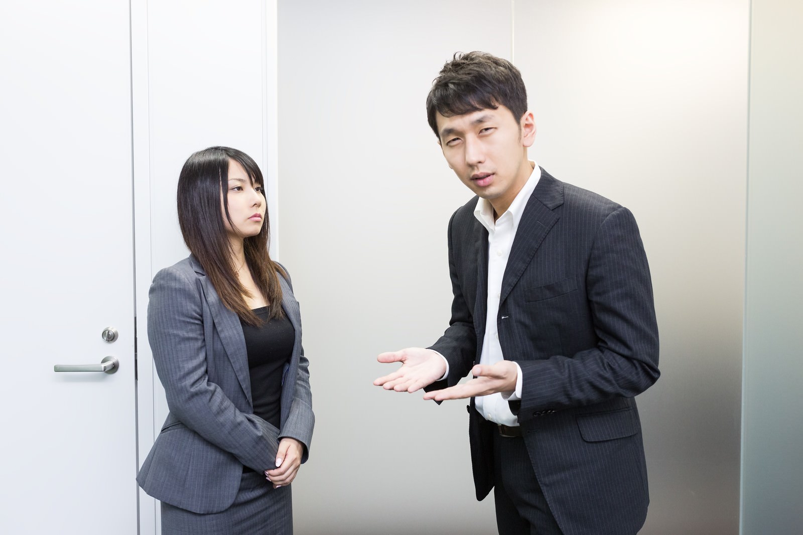 会議室で説明する紺色スーツの男性と、それを聞くグレースーツの女性［モデル：大川竜弥 Lala］