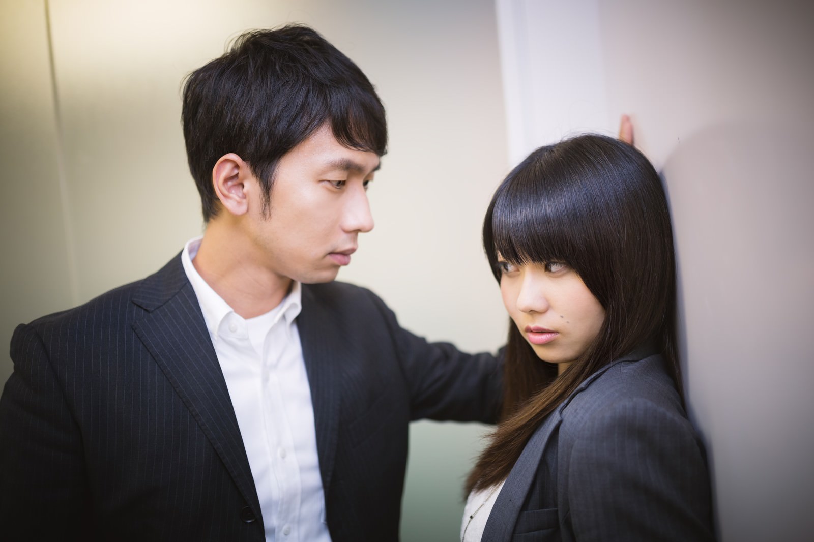 スーツ姿の男性上司が女性部下に壁ドンしている職場の一場面。女性は視線を逸らし拒否の表情を浮かべている［モデル：大川竜弥 Lala］