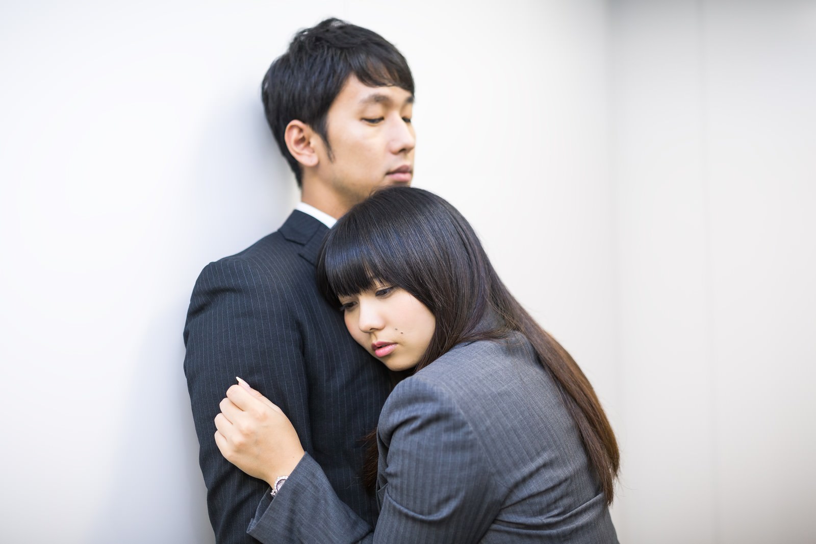 紺ストライプスーツの男性と灰色ジャケットの女性が白い壁に寄り添い、女性が男性に腕を絡めている場面［モデル：大川竜弥 Lala］