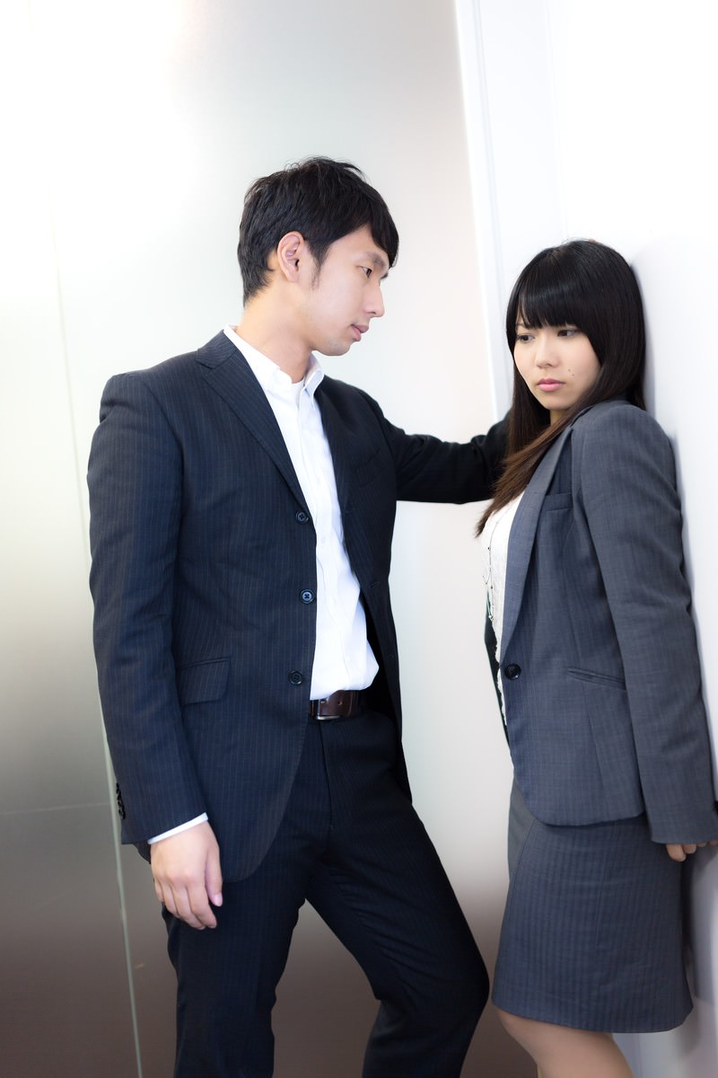黒いストライプスーツの男性がグレースーツの女性に壁ドンし、肩に手を置く職場シーン［モデル：大川竜弥 Lala］