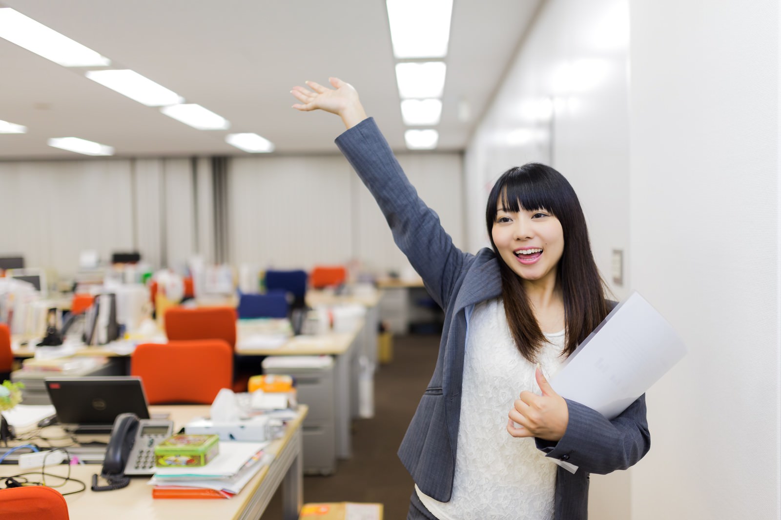 オフィスで資料を抱えながら手を振る笑顔の女性インターン生［モデル：Lala］