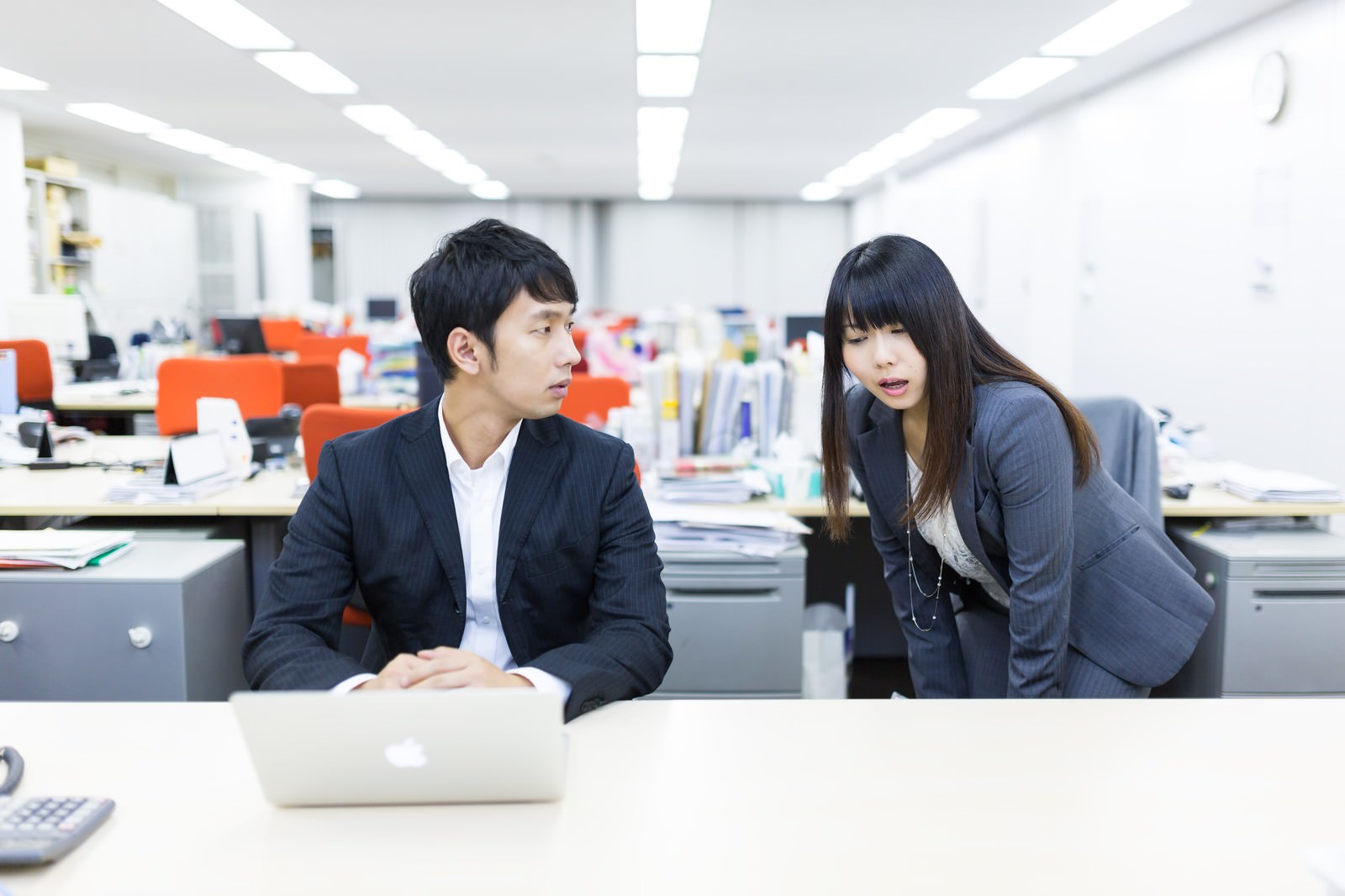 オフィスのデスク周辺で、紺スーツの女性社員が身を乗り出して話す様子を見つめる白シャツの男性上司［モデル：大川竜弥 Lala］