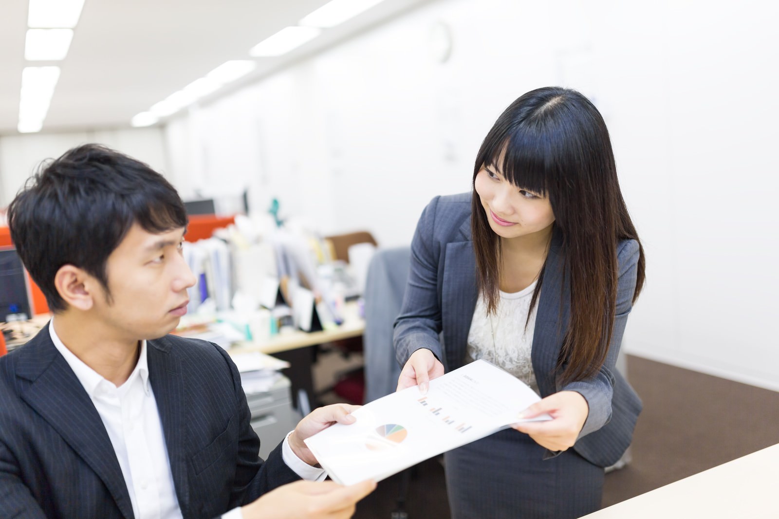 紺色スーツの男性がテーブルに座り資料を見つめる傍で、女性が立って資料を指し示す［モデル：大川竜弥 Lala］