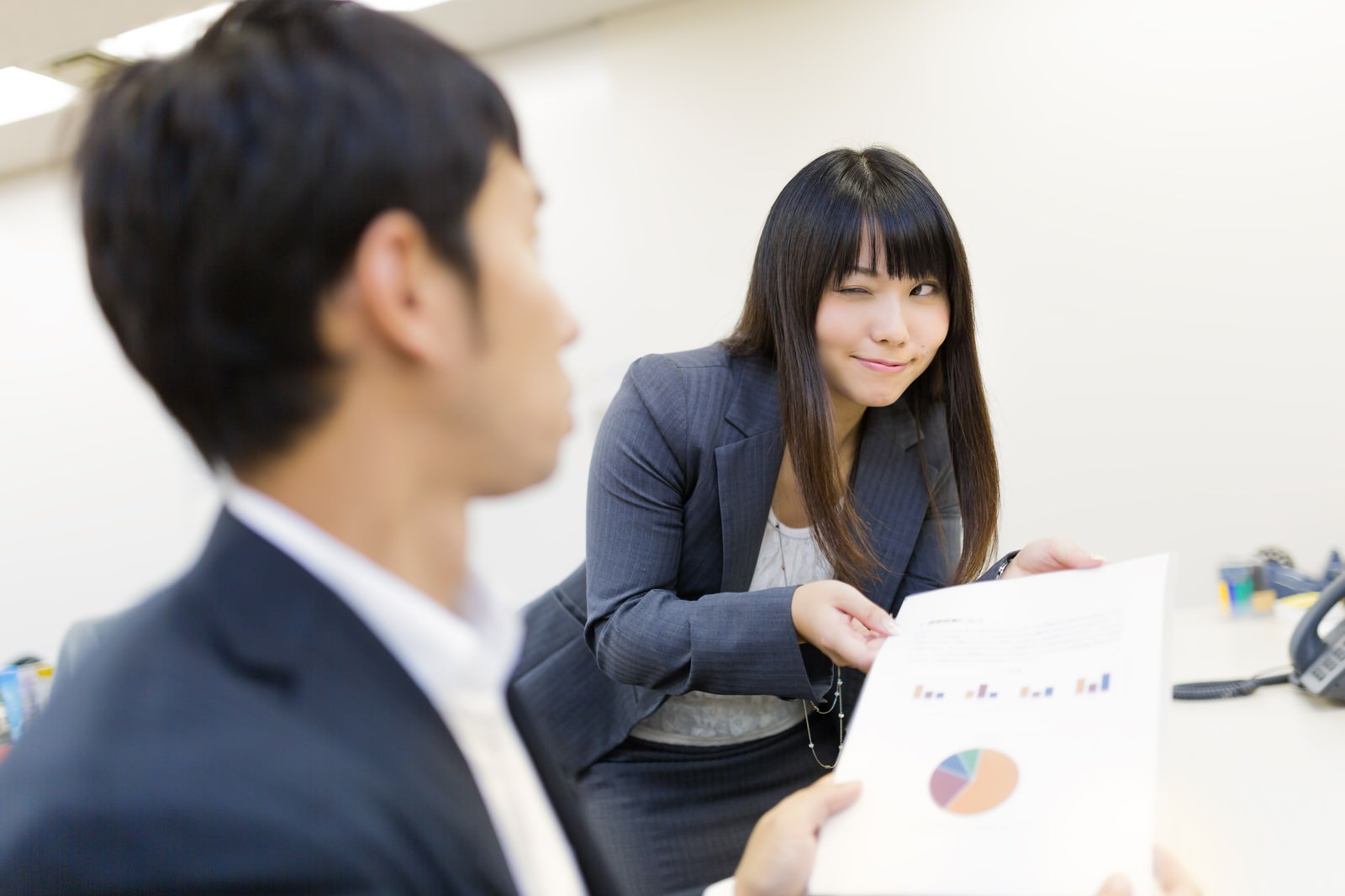 グラフ入り書類を手にウインクする濃紺スーツの女性社員と、話を聞く男性社員のオフィスシーン［モデル：大川竜弥 Lala］