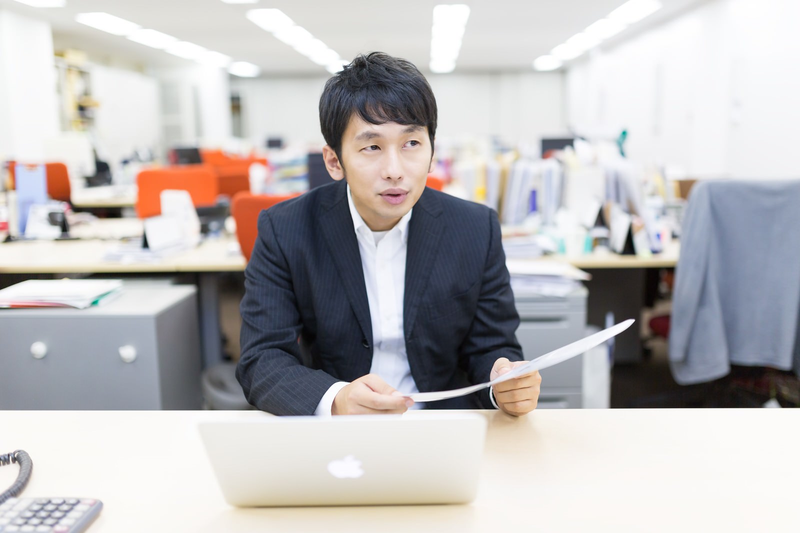 オフィスのテーブルで書類を持ちながら説明する紺スーツ姿の若い男性会社員［モデル：大川竜弥］