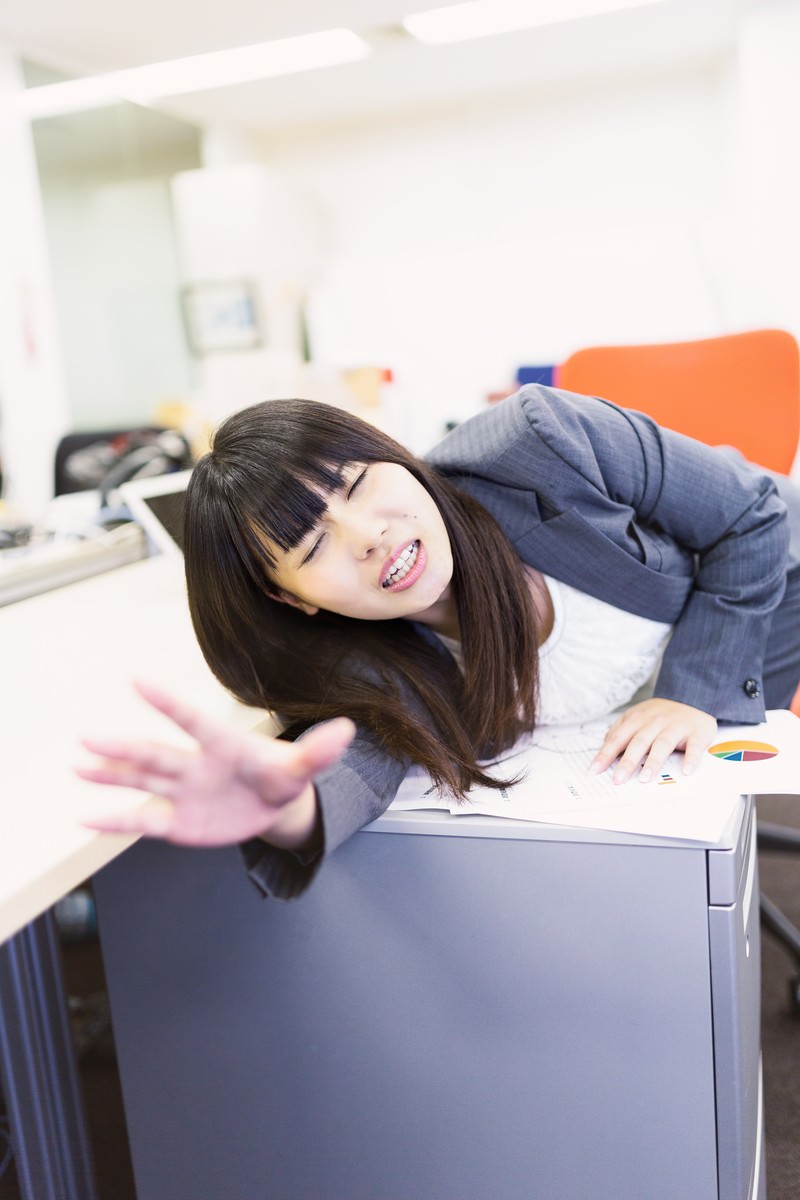 デスクで手を伸ばし、立ち去る同僚を呼び止める紺スーツの女性会社員［モデル：Lala］