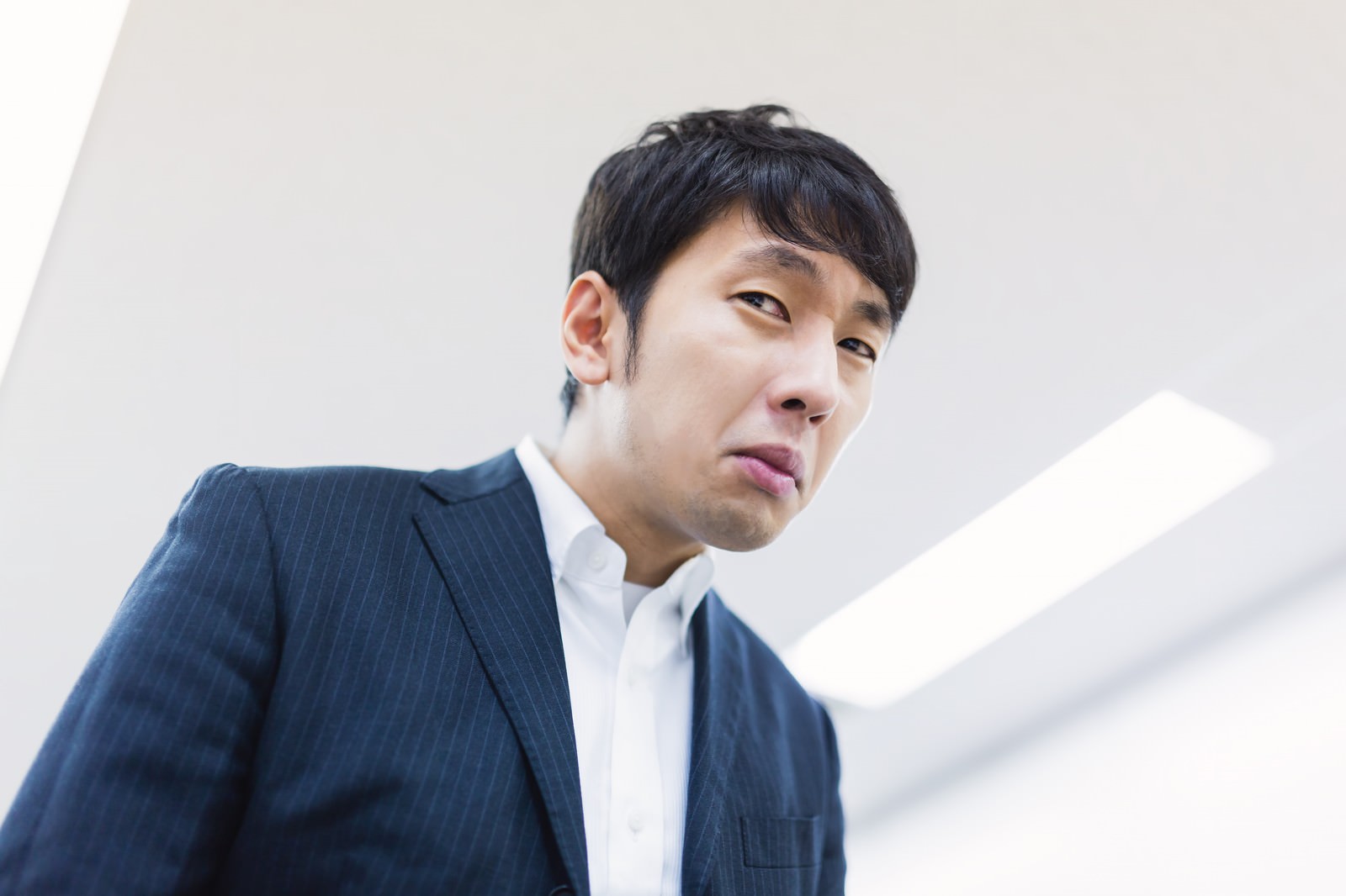 白い背景の前で濃紺スーツを着た男性が横向きに顔を上げ、口をへの字に結んだ困った表情をしている［モデル：大川竜弥］
