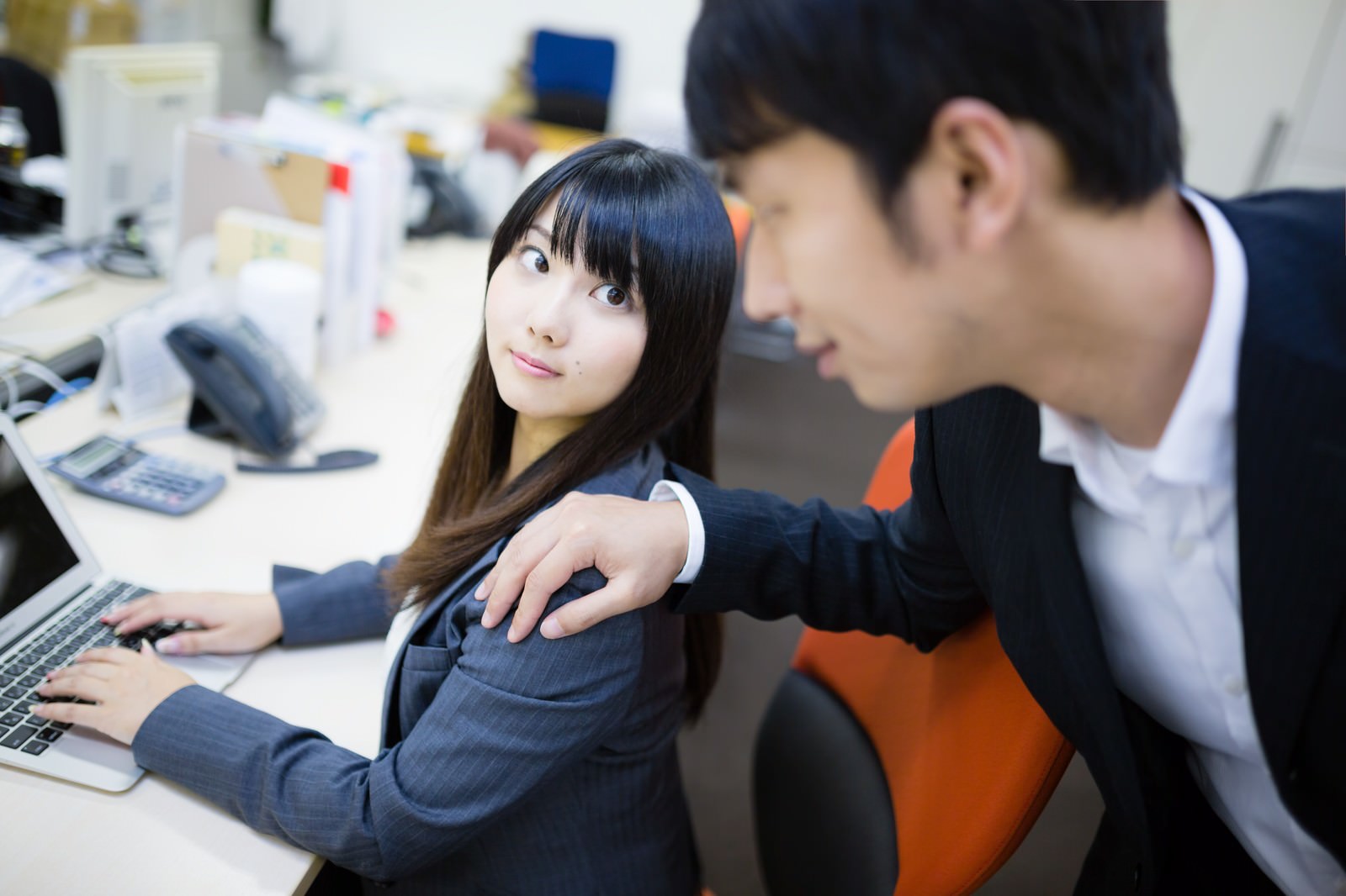 オフィスのデスクで、男性が女性の肩に手を置く中、振り返って不快感を表情で示す紺スーツの女性［モデル：大川竜弥 Lala］