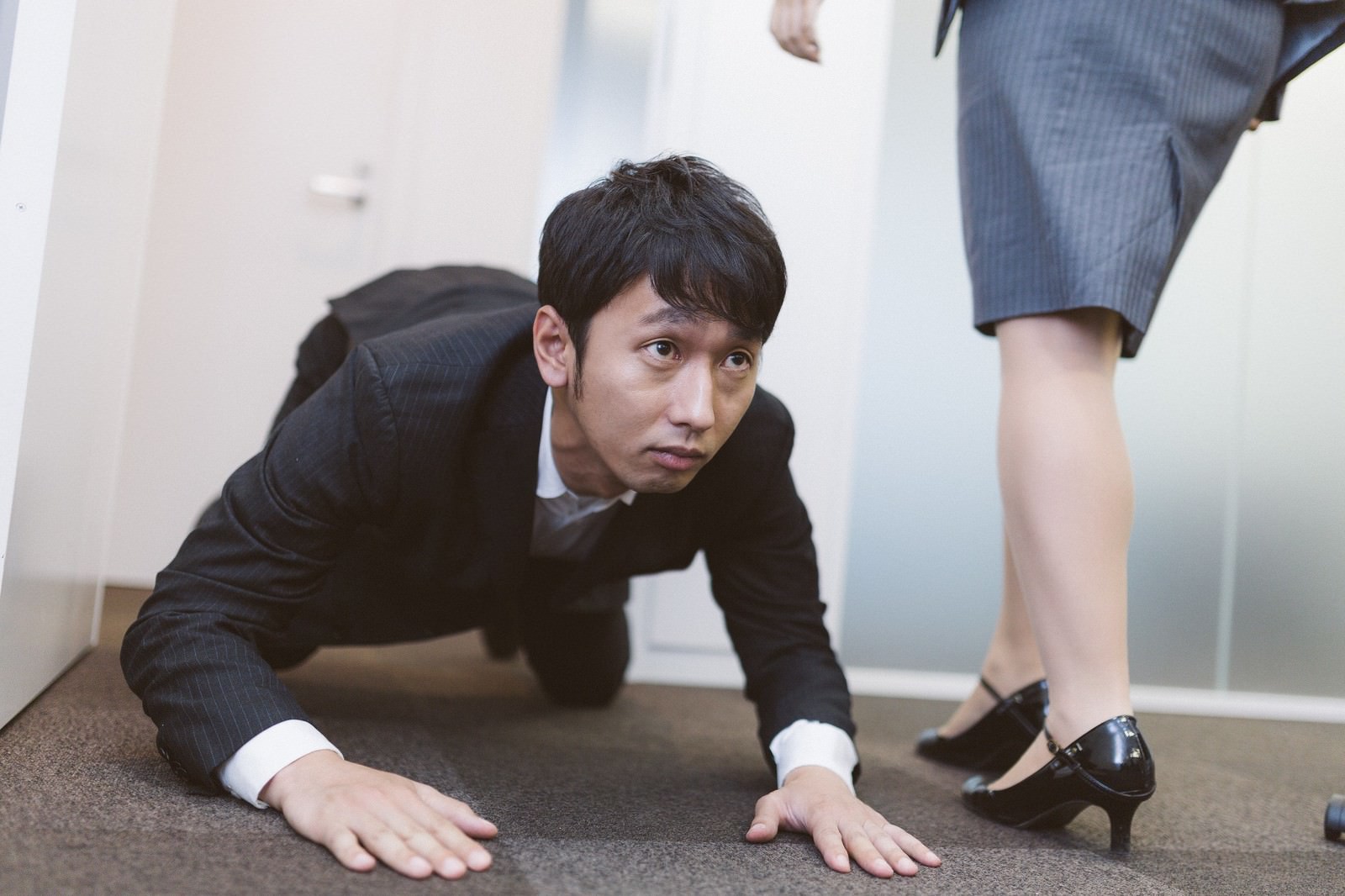 オフィスの床に両手をついて土下座する紺色スーツの男性が、立つビジネスウーマンに謝罪している［モデル：大川竜弥］