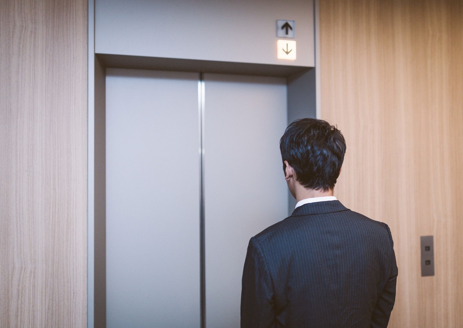 スーツを着た男性がエレベーターの前で昇降表示を見ながら待っている［モデル：大川竜弥］