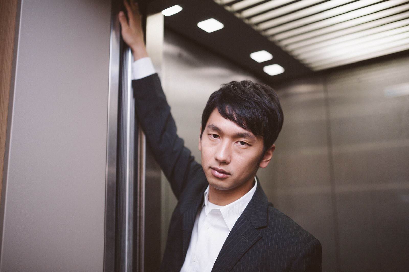 エレベーター内でスーツ姿の男性がドアの横に立ち、上を見ながらドヤ顔でポーズをとっている［モデル：大川竜弥］