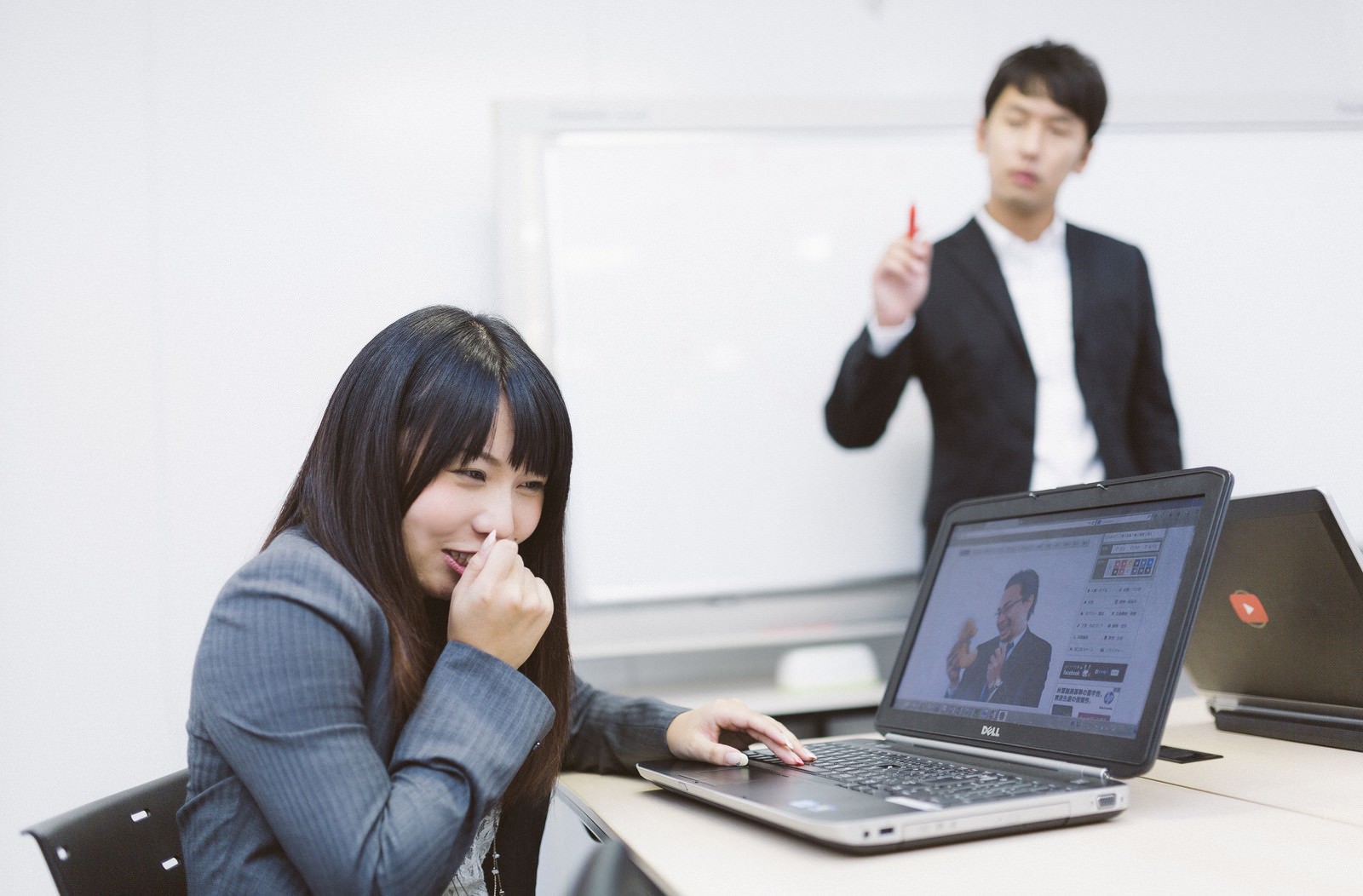 ホワイトボード前の会議室で、紺スーツの女性がパソコン画面を見つめ手を口に当て、黒スーツの男性講師が赤いペンで指導している［モデル：大川竜弥 Lala］