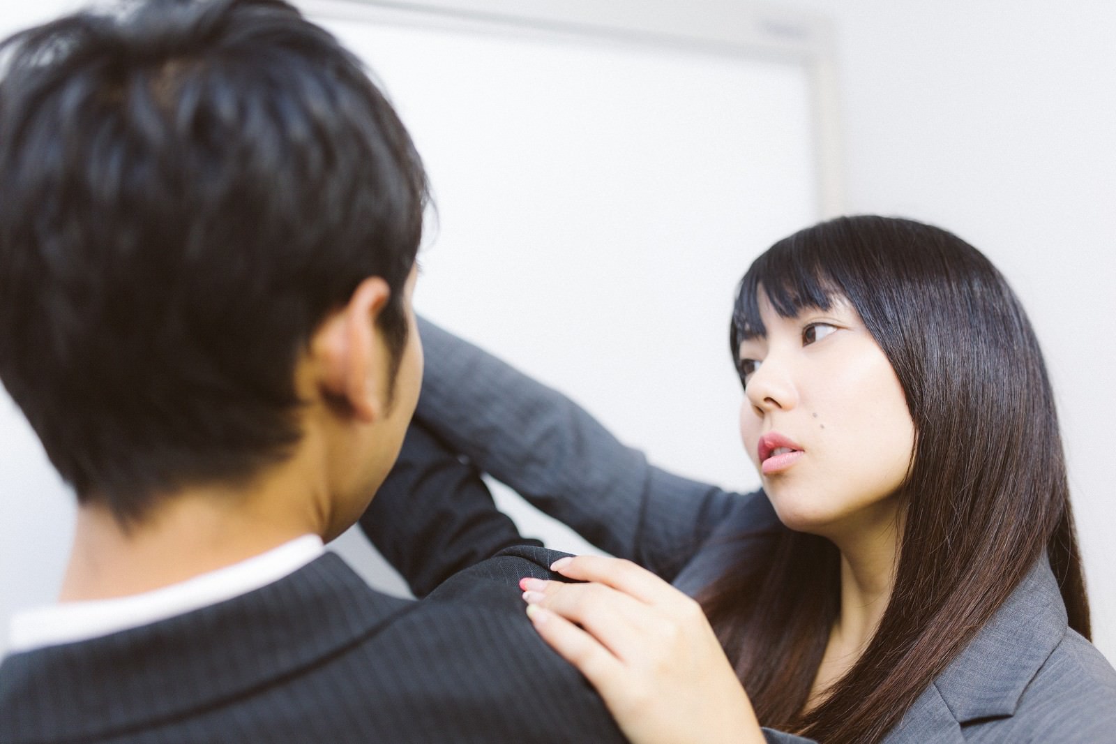 オフィスの白い壁を背景に、スーツの男性が女性にマンツーマンで丁寧に説明・指導している場面［モデル：大川竜弥 Lala］