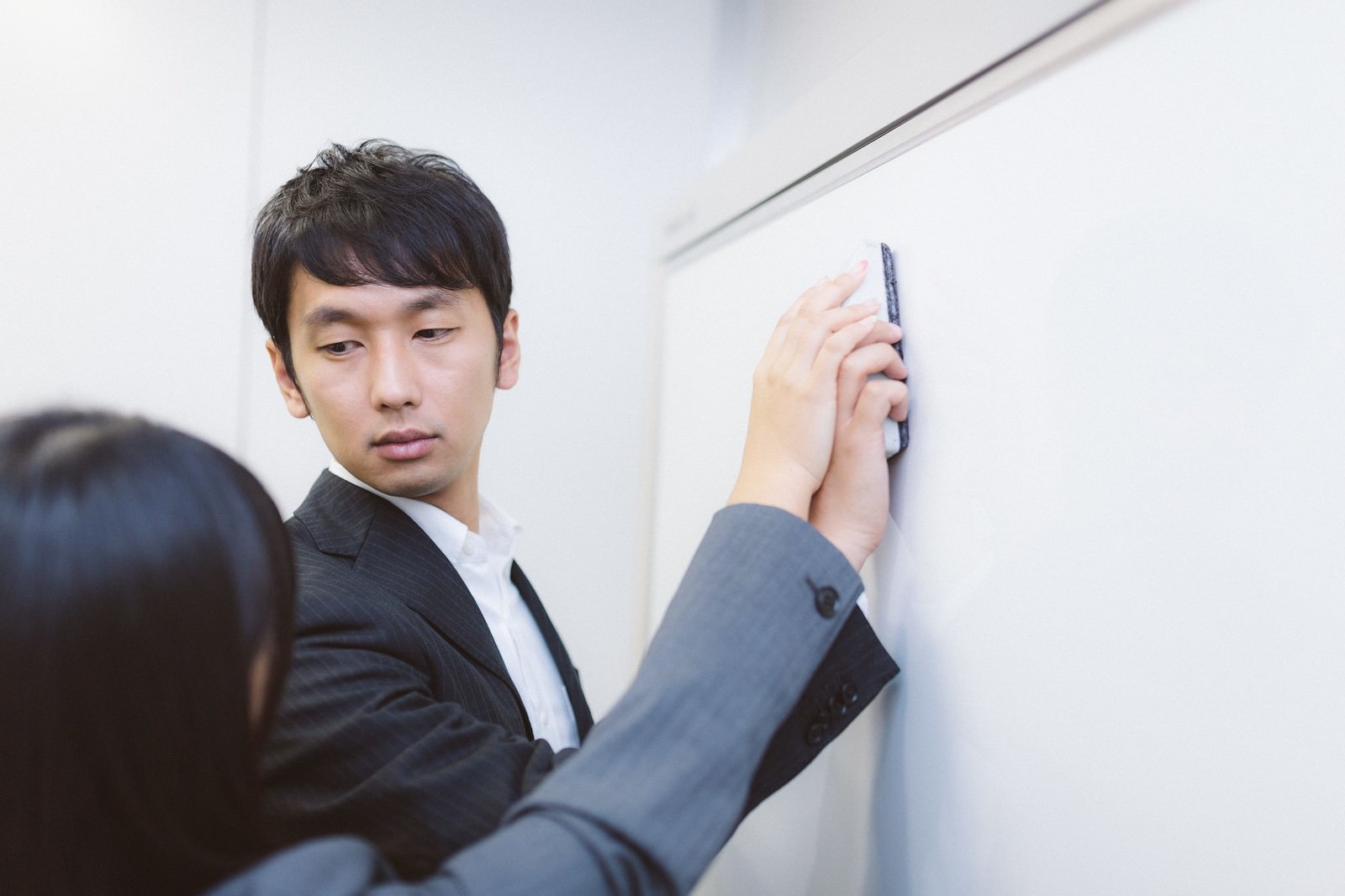 紺色スーツの男性がペンを持ってホワイトボードに向かい、女性の手が黒板消しを持って接近する職場研修の場面［モデル：大川竜弥 Lala］