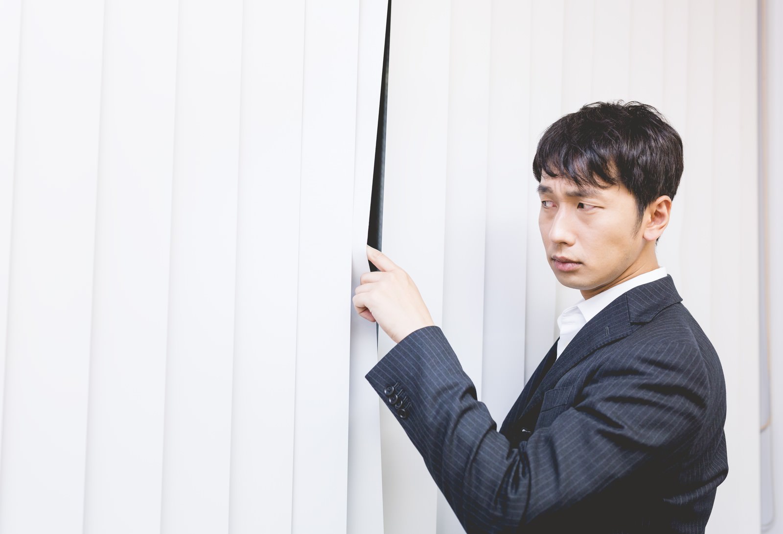 ブラインド越しに外を見つめるスーツ姿の男性会社員［モデル：大川竜弥］
