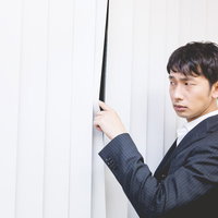 ブラインド越しに外の様子を気にする会社員男性の写真