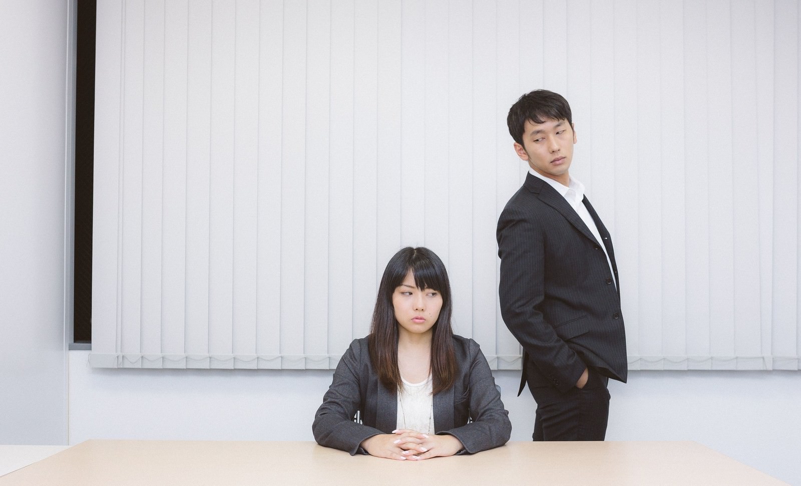 机を挟んで緊迫した面接場面。白シャツ女性と背後の黒スーツ男性が対峙［モデル：大川竜弥 Lala］