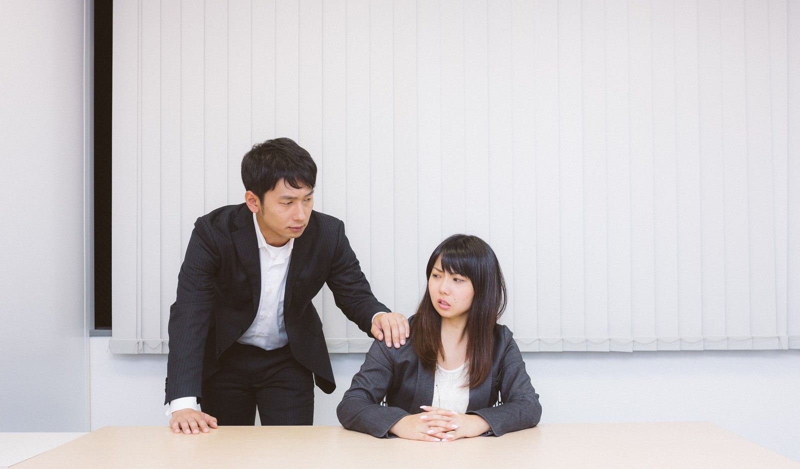 会議室で黒スーツの男性が濃紺スーツの女性に話しかけており、女性は視線を落とし困った表情で応じている［モデル：大川竜弥 Lala］