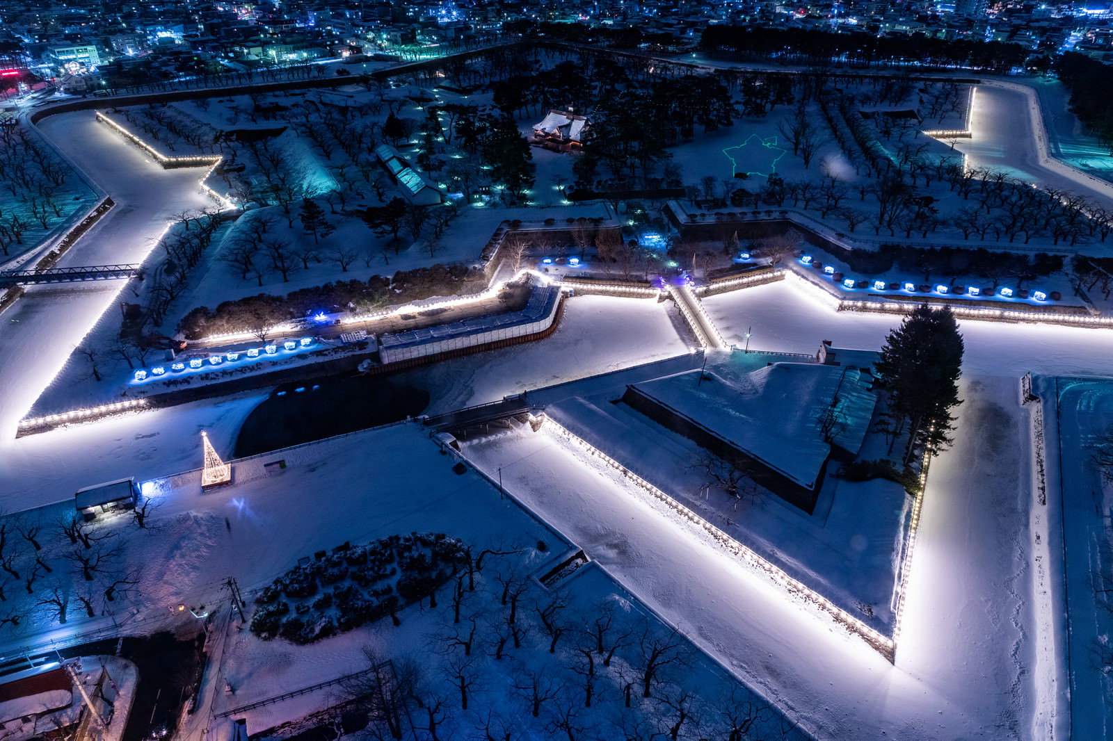 LED照明で縁取られた星型の五稜郭が雪に覆われ、背景に函館の街並みが見える夜間空撮