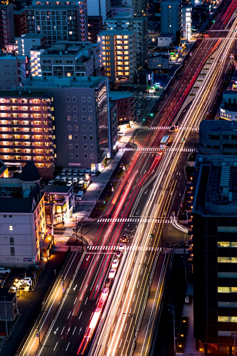夜の都市部で高層ビル群の間を走る道路に車のライトが描く光跡