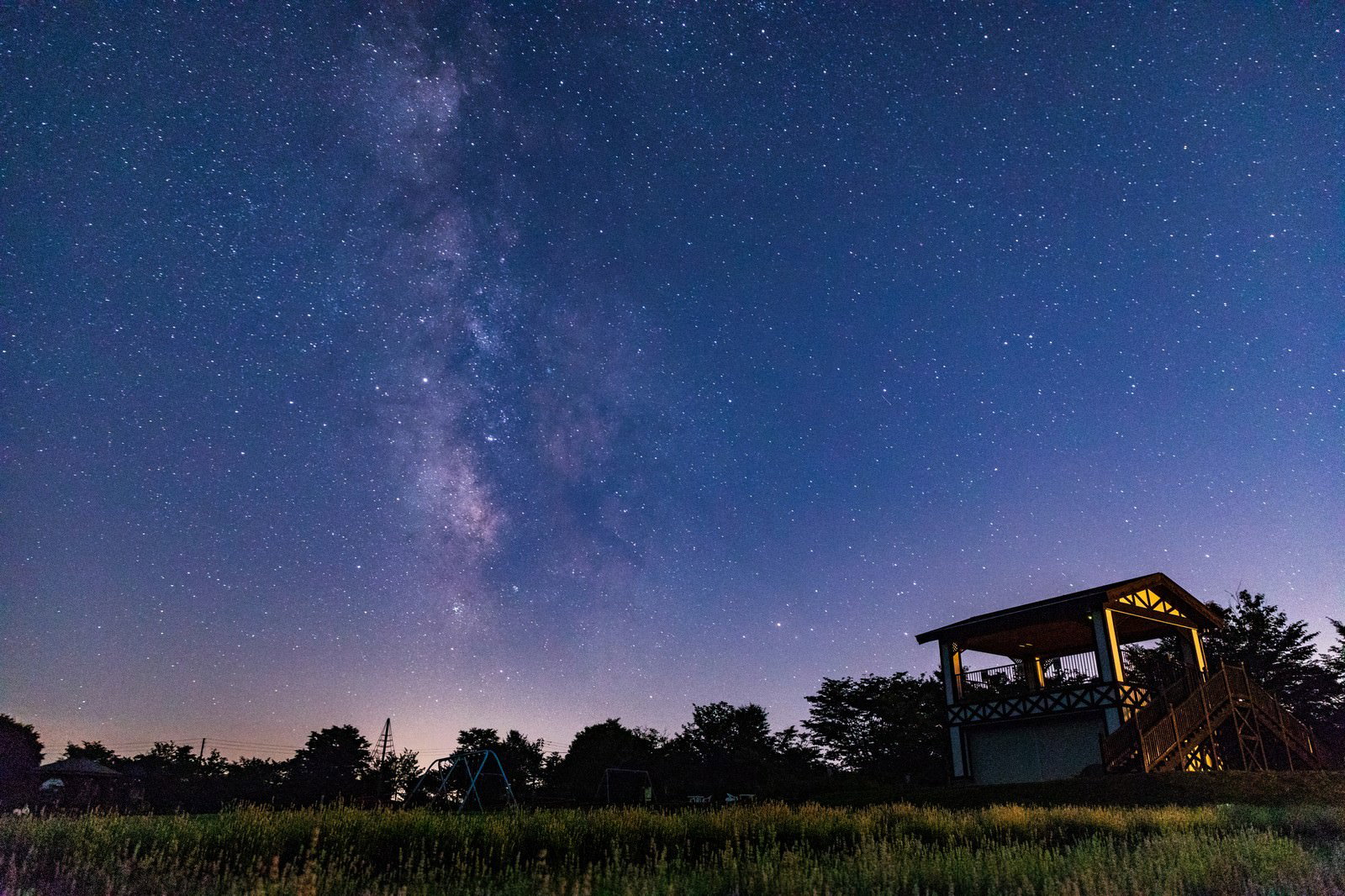 深い青色の夜空に天の川が浮かび、手前にラベンダー園と観測小屋がある夜景