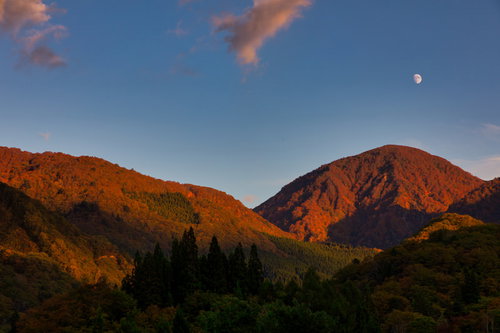 夕暮れに染まる紅葉した山と空に浮かぶ月の秋風景
