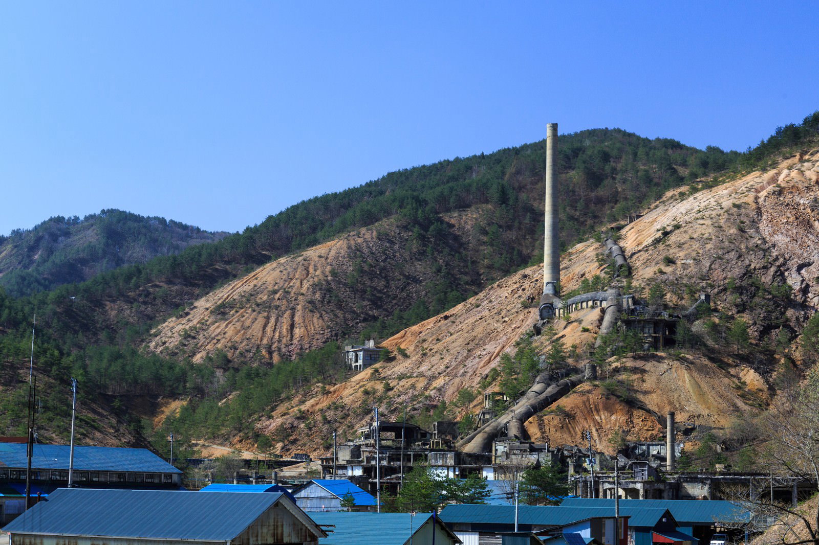 荒れた山肌に立つ鉱山跡の煙突と建物、森に囲まれた採掘遺跡