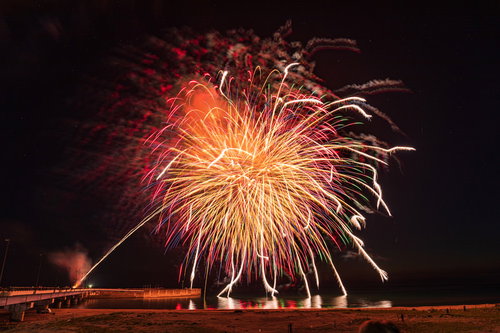 海に打ち上がる花火のオレンジと赤い光跡が夜空を彩る