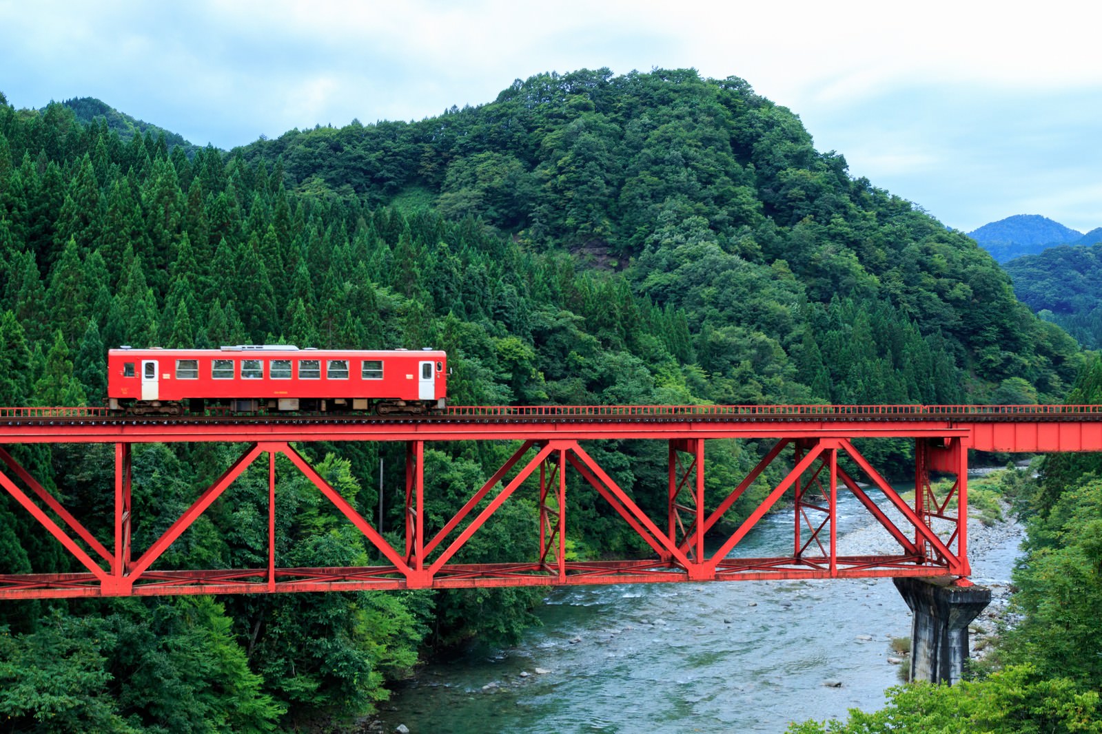 赤いトラス橋を走行する秋田内陸鉄道の赤い電車と、両側の深い緑の山林
