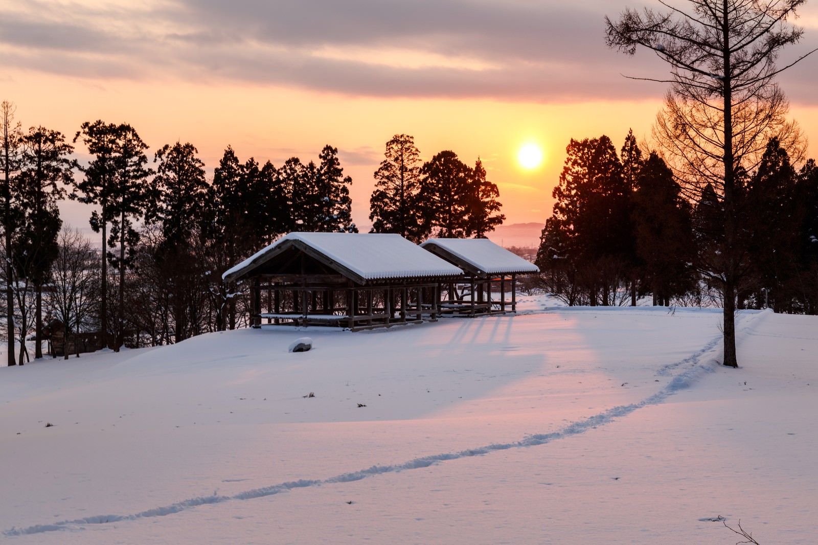 雪に覆われた田舎の原っぱに佇む茅葺き屋根の建物と夕日を見守る樹木たちの冬景色