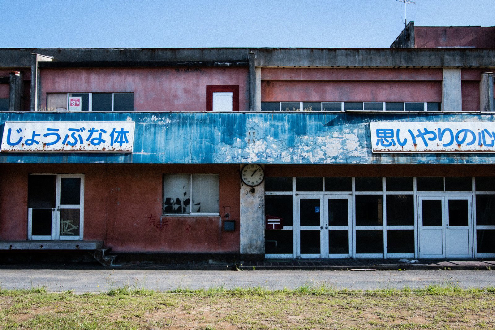 赤い廃校の校舎。青い錆びた看板と古い時計がある