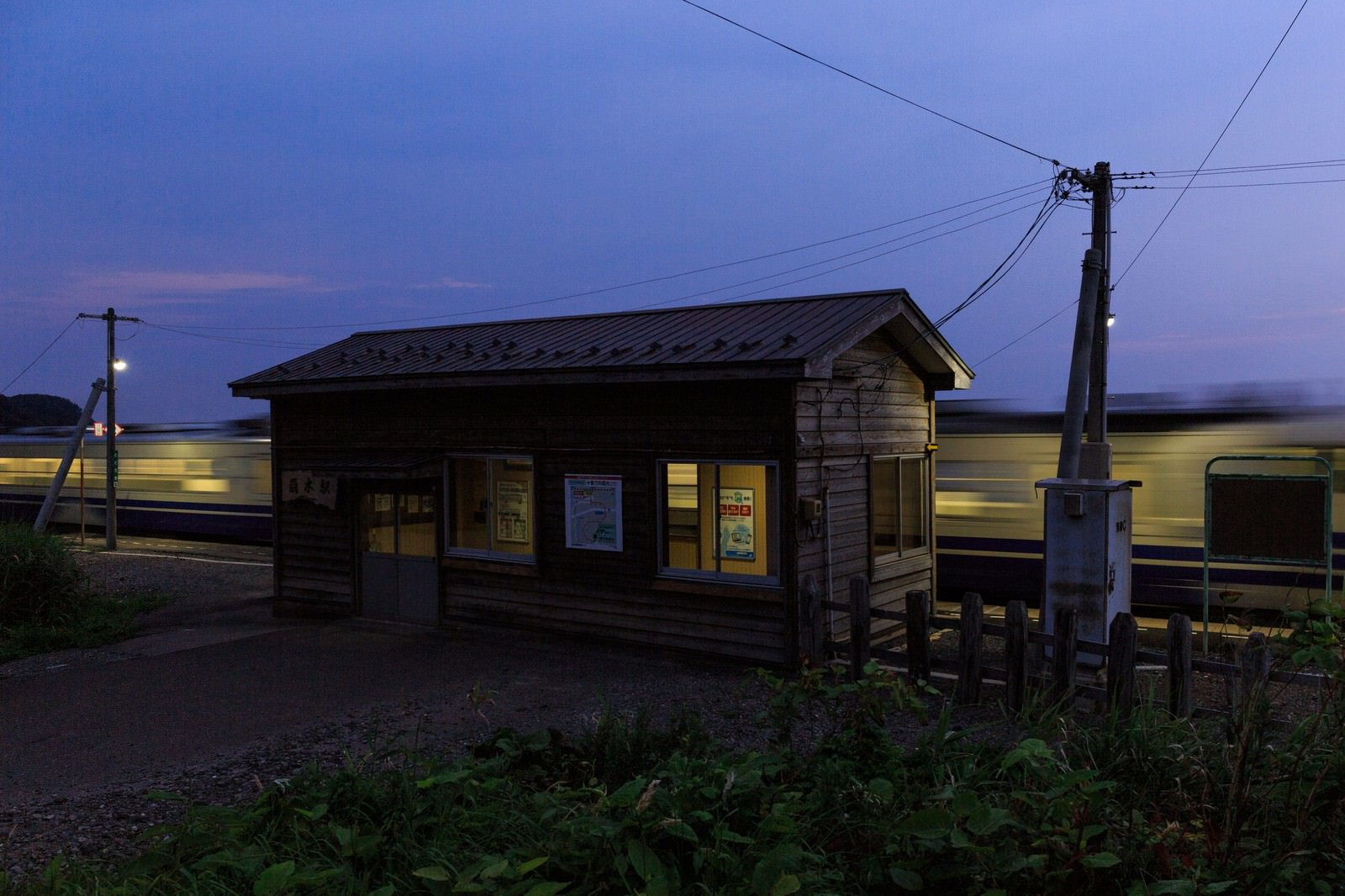夕暮れに照明が灯る木造の無人駅とローカル線の電車