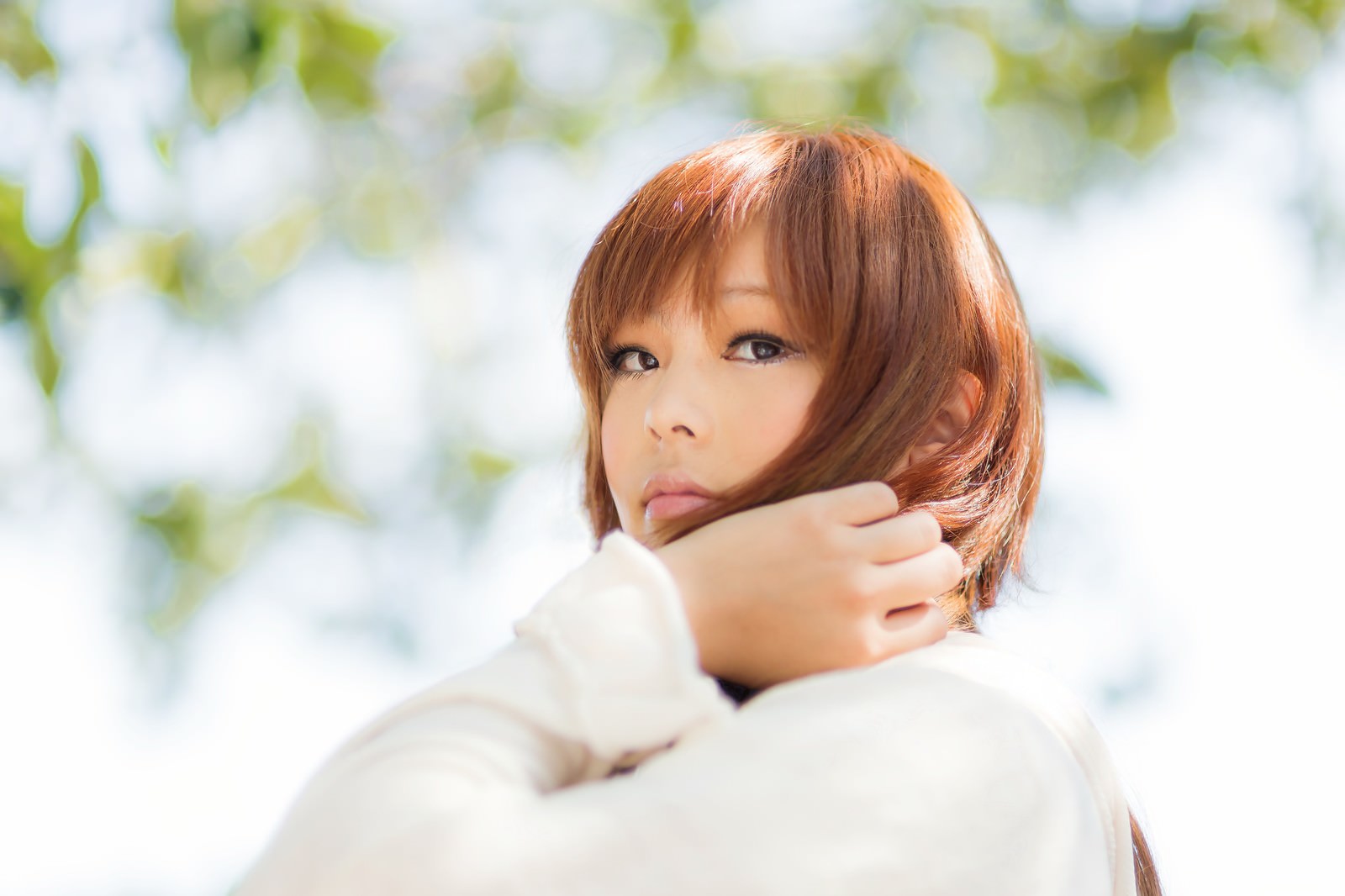 新緑の背景で振り返る白いトップスを着た茶髪女性［モデル：あみ］